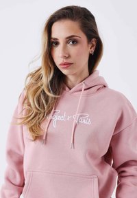 Sudadera con capucha rosa con un bolsillo frontal, con texto en cursiva blanca que dice "Project x Paris". Tela suave, cordones y extremos plateados.