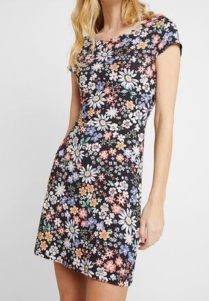 Femme portant une robe noire à manches courtes ornée de motifs de pâquerettes multicolores et de petites fleurs, debout devant un fond uni.