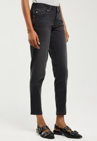 Osoba stojící na jednobarevném pozadí, oblečená v úzkých černých džínách a černých slingback botách s ostrou špičkou a stříbrnými přezkami.