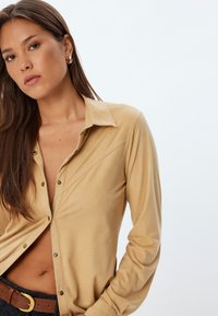 Beige skjorte med knapper og en blød tekstur, der har en krave og skrå sømdetaljer. Stylet med et brunt bælte og mørke jeans.