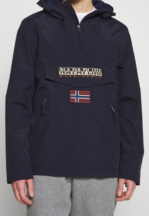 Marineblaue Napapijri Kapuzenjacke mit Frontreißverschluss, Klappentasche, norwegischem Flaggen-Patch, getragen mit hellgrauen Hosen.