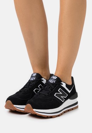 Gambe con indosso sneakers New Balance 574 nere con dettagli bianchi e suole color gomma, in piedi su una superficie bianca.