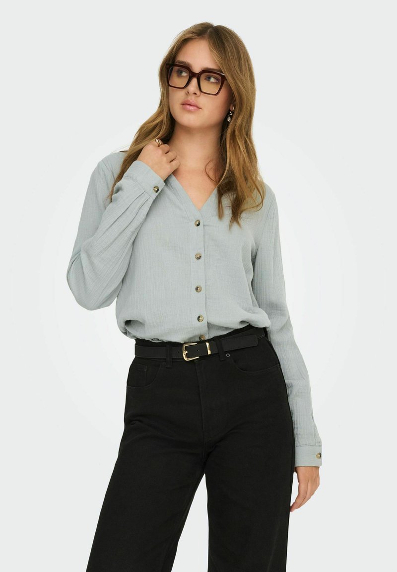 Femme portant de grandes lunettes marron, chemisier gris clair boutonné, pantalon noir avec ceinture, posant avec la main touchant le col, regardant sur le côté.