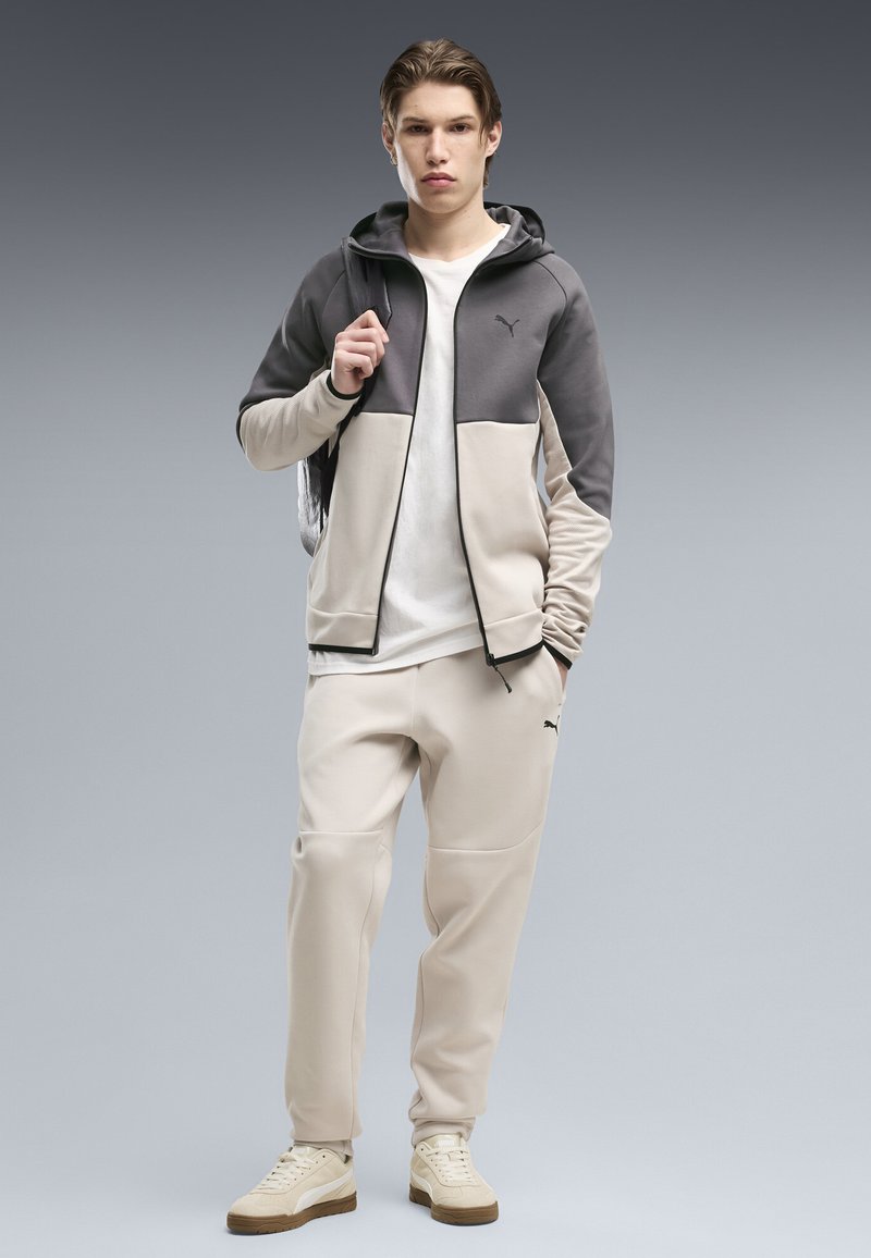 Jeune homme portant un survêtement Puma beige et gris et des baskets crème, debout, une main dans la poche et l'autre tenant la sangle d'un sac à dos.