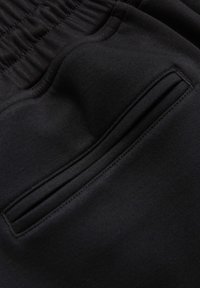 Gros plan d'un pantalon en tissu noir montrant une taille élastique et une poche fendue verticale avec détails de couture.