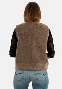 Persona con capelli castani lisci, che indossa un gilet di pile marrone sopra una maglietta in rete nera floreale e jeans blu, vista da dietro.