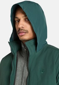 Grüne, mit Reißverschluss versehene Jacke mit Kapuze, die eine strukturierte graue Innenschicht und ein gesticktes Logo auf der Brust hat. Leichtes Material.