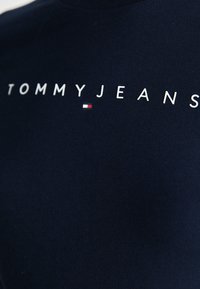 Tengerszínű pamut póló fehér betűkkel, amelyen a "TOMMYJEANS" felirat és egy kis piros-fehér logó található. Kerek nyakú, rövid ujjú, sima tapintású.