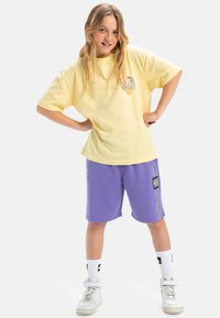 Geel katoenen T-shirt met een grafische print, gecombineerd met paarse katoenen shorts met zijaccenten en een label, afgewerkt met witte sneakers.