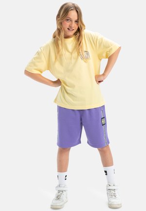 T-shirt en coton jaune avec un imprimé graphique, associé à un short en coton violet avec des accents sur les côtés et une étiquette, le tout complété par des baskets blanches.