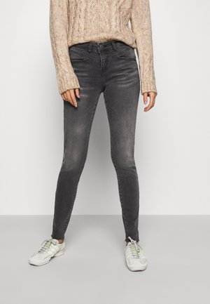 Skinny jeans in donkergrijs denim met vervaagde accenten, gecombineerd met een beige kabelgebreide trui en witte sneakers met felgroene veters.