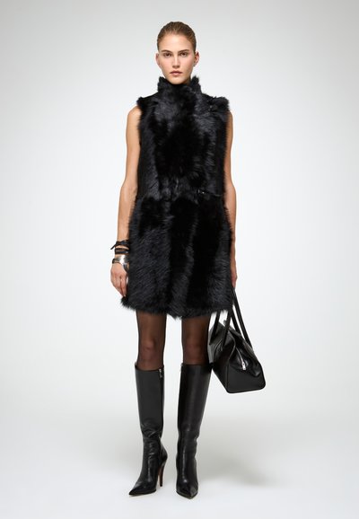 Juoda kailinė liemenė su aukštu apykakle, užsegta mygtuku, vilkima ant permatomų juodų pėdkelnių ir juodų aukštakulnių batų iki kelių. Papuošta juoda rankine.