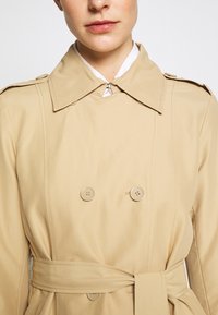 Abrigo trench beige con diseño de doble botonadura, cuello grande, dos botones y cinturón de cintura atado, hecho de tela lisa.