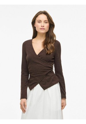 Moteris su ilgais rudos spalvos plaukais, dėvinti aptemptą tamsiai rudos spalvos užrišamą megztinį ir baltą sijoną, stovinti prie paprasto balto fono.