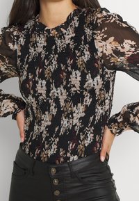 Blouse noire à motifs floraux avec un corps froncé et des manches bouffantes transparentes. Elle présente des teintes de beige, bordeaux et vert dans un motif abstrait.