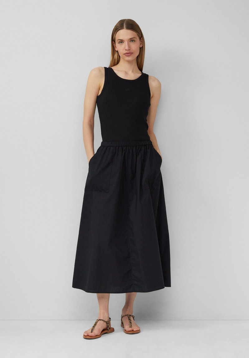 Schwarzes Tanktop und langer schwarzer Rock mit elastischem Bund, ausgestattet mit Seitentaschen. Sandalen mit einem Leopardenmuster-Riemen. Glatte Textur.