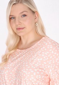 Světle růžový top s bílými nepravidelnými puntíky, kulatým výstřihem a krátkými rukávy. Model má dlouhé blonďaté vlasy a kruhové náušnice.