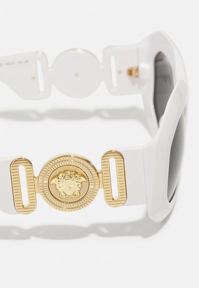 White sunglasses featuring a chunky, glossy frame with dark lenses, accented by a gold round medallion with a raised design.