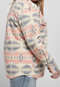 Patterned jacket featuring a geometric design in pink, blue, and cream. Made of fabric with a slightly textured finish. Buttoned cuffs.