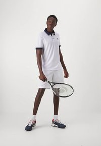 Hvid polo med navy krave, kombineret med hvide shorts. Modellen holder en sort tennisketsjer og har navy og røde tennissko på.