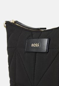 Černá prošívaná taška z textilu se hladkou koženou etiketou s vyraženým nápisem "BOSS" zlatou barvou. Design obsahuje úhlové prošívání a zlatý zip.