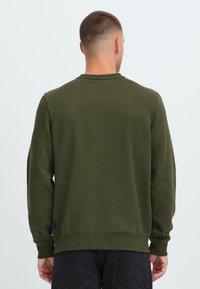 Mann med kort hår iført en enkel olivengrønn sweatshirt og svarte bukser, stående med ryggen til kameraet mot en ensfarget bakgrunn.