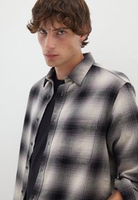 Camisa a cuadros en negro y crema con botones en el frente, un bolsillo y cuello tipo camisa. Se lleva sobre una camiseta negra, hecha de tejido texturizado.