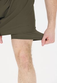 Olivgrüne Sportshorts mit glatter Textur, ausgestattet mit einem seitlichen Schlitz und dem "ENDURANCE"-Logo am Saum. Eine Hand greift den Stoff.