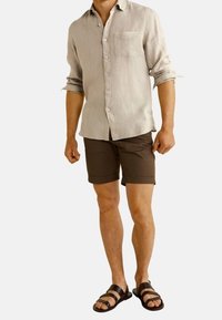 Chemise en lin beige clair avec une poche, short marron et sandales noires. La chemise est à boutons pression et les manches sont retroussées.