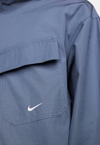 Camisa Nike de tejido azul marino con patrón texturizado, que presenta un bolsillo con solapa y un acento de logo blanco. Diseño ligero y transpirable.