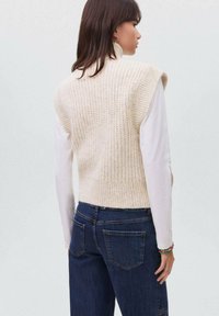 Maglione senza maniche in crema con un motivo testurizzato, indossato su una camicia bianca a maniche lunghe, abbinato a jeans blu scuro.