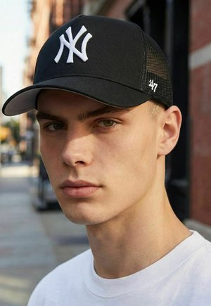 Junger Mann mit schwarzer New York Yankees Baseballkappe und weißem Hemd, der draußen in der Nähe eines Backsteingebäudes steht.