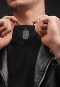 Ciondolo in metallo a forma di medaglietta su una catena nera, caratterizzato da un logo e un'incisione. Indossato sopra una maglietta nera, sostenuto da una persona. Texture liscia.