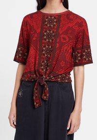 Röd mönstrad topp med korta ärmar, en knytframsida och intrikata blommönster i gult och svart, kombinerad med mörka löst sittande byxor.