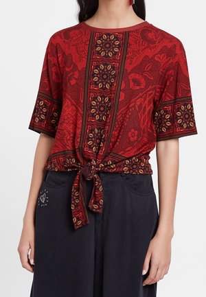 Haut à motifs rouges avec des manches courtes, un devant noué et des dessins floraux complexes en jaune et noir, associé à un bas ample et sombre.