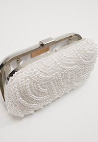 Vit kuvertväska med en texturerad pärldesign, en silvertonad lås och en rundad form. Har detaljerat pärlarbete som bildar vågmönster.