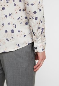 Lightweight, floral-patterned shirt with cream background, purple and red accents, and buttoned cuffs. Paired with gray textured trousers.