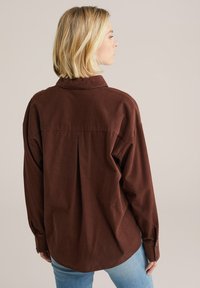 Brown cotton shirt with a relaxed fit, long sleeves, shirt collar, and a pleat detail at the back. Worn over light blue jeans.