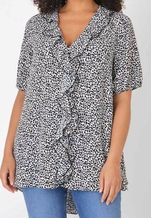 Kobieta nosząca białą bluzkę z krótkim rękawem, z czarnym nieregularnym wzorem kropek i falbanami z przodu, w połączeniu z niebieskimi jeansami.