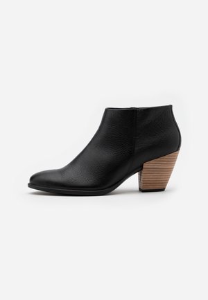 Bottine en cuir noir avec un bout pointu et un talon bloc en bois empilé sur fond blanc.