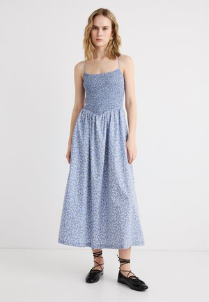 Junge Frau trägt ein blaues Midikleid mit floralem Muster, dünnen Trägern, einem gerafften Oberteil und schwarzen Ballerinas mit Schnürung, steht vor weißem Hintergrund.