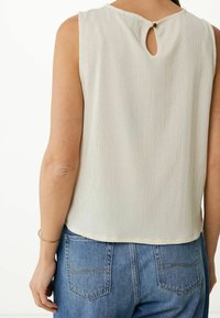 Haut sans manches beige clair avec une surface texturée, détail dos avec ouverture en forme de goutte, et ourlet arrondi, associé à un jean bleu.