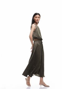 Olijfgroen, mouwloze tweedelige outfit van textuurstof; crop top gecombineerd met een hoge taille rok en witte hakken.