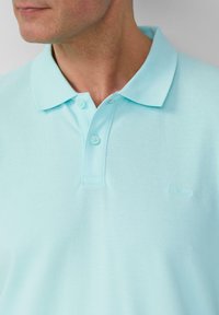 Man in een lichtblauw polo shirt met twee vastgemaakte knopen, zichtbare kraag en een subtiel merkembleem op de borst.