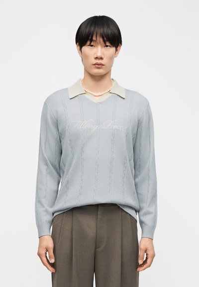 Νεαρός άνδρας με μαύρα μαλλιά που φοράει ανοιχτό μπλε πουλόβερ με την επιγραφή «Filling Pieces», μπεζ πουκάμισο με γιακά, περιδέραιο από μαργαριτάρια και καφέ παντελόνι.