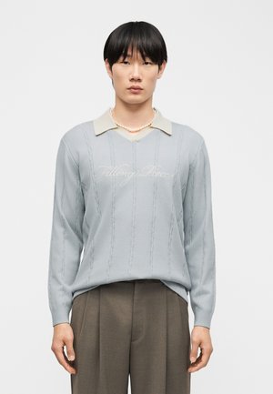 Νεαρός άνδρας με μαύρα μαλλιά που φοράει ανοιχτό μπλε πουλόβερ με την επιγραφή «Filling Pieces», μπεζ πουκάμισο με γιακά, περιδέραιο από μαργαριτάρια και καφέ παντελόνι.