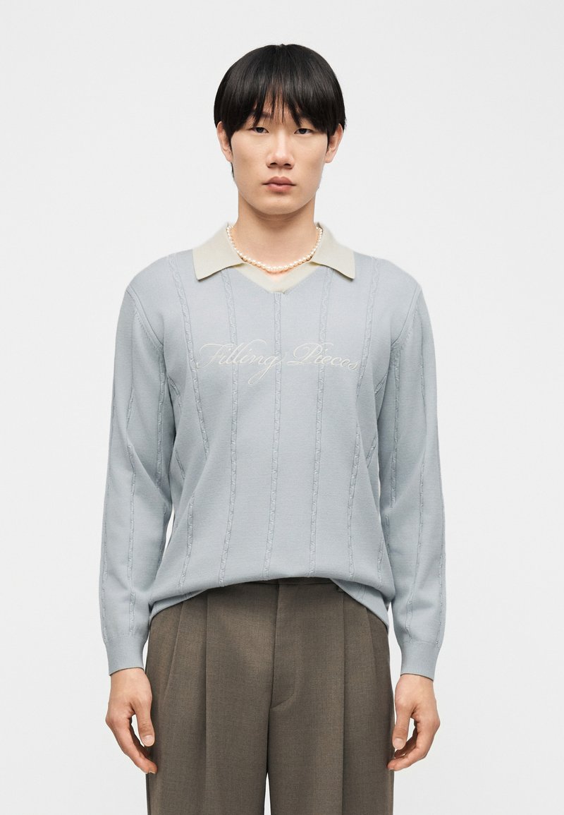 Mladý muž s černými vlasy, který má na sobě světle modrý svetr s nápisem "Filling Pieces", béžovou košili s límečkem, perlový náhrdelník a hnědé kalhoty.