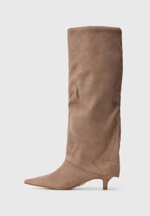 Bottes en suede marron hautes jusqu'au genou, à bout pointu et petit talon de type kitten sur fond blanc uni.