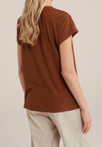 Brun t-shirt med korta ärmar och ett strukturerat zigzag-mönster, rund halsringning och avslappnad passform, stylad med ljusa byxor.