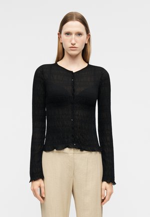 Donna che indossa un top nero attillato con maniche lunghe e bottoni, caratterizzato da un motivo testurizzato sottile, e pantaloni beige a vita alta, in piedi contro uno sfondo semplice.